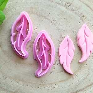 May include: Four pink plastic cookie cutters shaped like feathers. The cutters are on a light brown wooden surface.