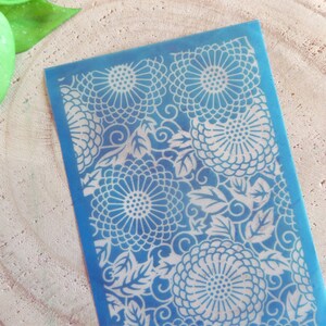 Può includere: Uno stencil blu con un motivo floreale di grandi fiori e foglie. Lo stencil è rettangolare con un bordo. Il disegno è bianco su sfondo blu. Probabilmente viene utilizzato per l'artigianato o per scopi decorativi, come la pittura.