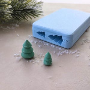 May include: A light blue silicone mold with two Christmas tree shapes. Two green Christmas tree-shaped decorations are next to the mold.