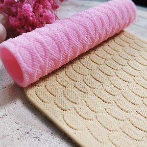 Può includere: Un mattarello in silicone rosa con una superficie testurizzata e un tappetino in silicone beige con un motivo ondulato. Il mattarello è appoggiato sul tappetino.