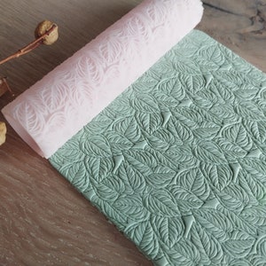 May include: A pink silicone rolling pin with a leaf pattern is rolling over a green fondant sheet with a leaf pattern.