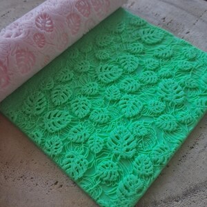 Può includere: Un mattarello cilindrico rosa chiaro con un motivo a foglie di monstera in rilievo, posizionato sopra un foglio quadrato di materiale verde brillante impresso con lo stesso dettagliato disegno di foglie di monstera su tutta la superficie.