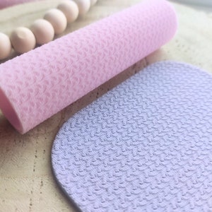 Puede incluir: Primer plano de dos hojas de tela texturizada. Una hoja es de color rosa claro y está enrollada en forma de cilindro. La otra hoja es de color morado claro y está plana. Ambas hojas tienen un patrón texturizado de pequeños puntos en relieve.