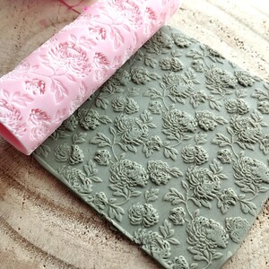 Può includere: Un mattarello rosa con motivo floreale è appoggiato accanto a un tappetino grigio strutturato, anch'esso con un motivo floreale. Entrambi gli articoli presentano un motivo a crisantemo in rilievo, ideale per progetti di panificazione e artigianato.