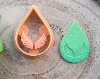 Peut inclure: Un emporte-pièce en forme de goutte d'eau et un objet vert en forme de goutte d'eau avec des motifs d'ailes. L'emporte-pièce est de couleur pêche et présente un motif d'ailes à l'intérieur de la forme de goutte d'eau.