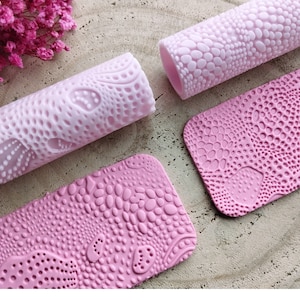 Peut inclure: Quatre rouleaux à pâtisserie et tapis en silicone rose avec un motif texturé de petits cercles et ovales. Les rouleaux à pâtisserie sont cylindriques et les tapis sont rectangulaires.