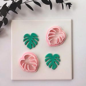 Puede incluir: Dos cortadores de galletas de plástico rosa claro con forma de hoja de monstera y dos formas de hoja de monstera verdes texturizadas hechas de arcilla o masa, dispuestas en cuadrado sobre una superficie blanca.