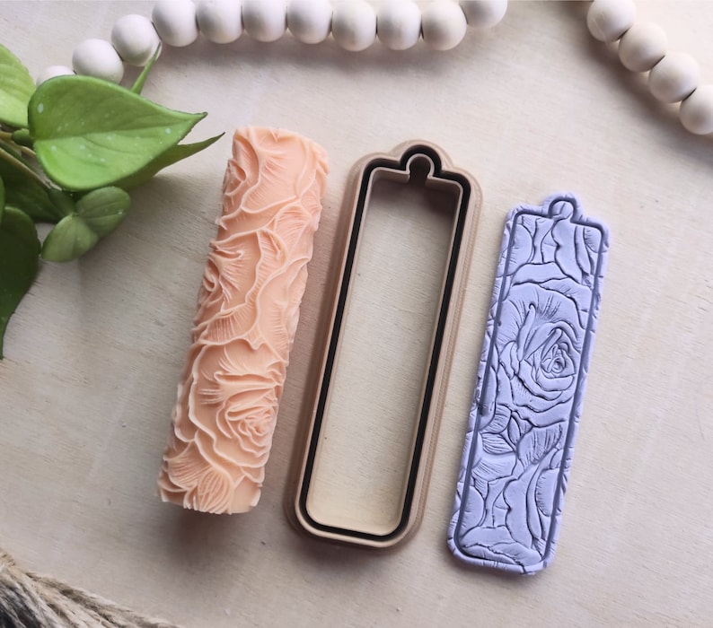 Op de afbeelding: Een set van drie polymer clay gereedschappen voor het knutselen. De gereedschappen zijn een deegroller met een rozenpatroon, een rechthoekige uitsteker en een rechthoekige stempel met een rozenpatroon. De gereedschappen liggen op een licht houten oppervlak.