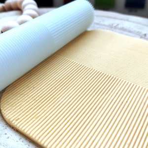 Op de afbeelding: Een lichtblauwe deegroller met een getextureerd oppervlak rolt over een lichtbeige getextureerd oppervlak. Het oppervlak heeft een patroon van parallelle lijnen.