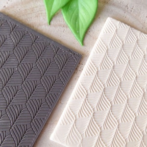 Può includere: Due tappetini da forno in silicone con un motivo a foglie. Il tappetino a sinistra è grigio scuro e il tappetino a destra è beige chiaro.