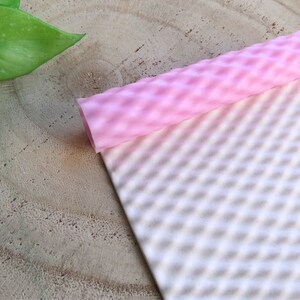 Puede incluir: Un rodillo texturizado rosa y una estera de silicona texturizada blanca. Ambos tienen un patrón de diamante.