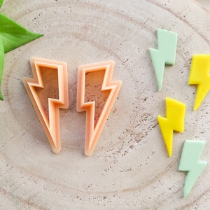 Può includere: Due tagliabiscotti a forma di fulmine in plastica rosa su una superficie di legno con cinque biscotti a forma di fulmine colorati.