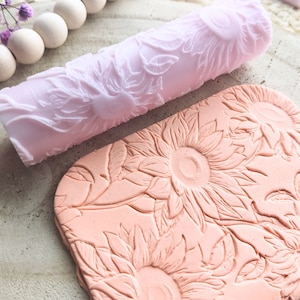 Può includere: Un mattarello rosa con un motivo a girasole è mostrato accanto a un pezzo di argilla con lo stesso motivo a girasole in rilievo.