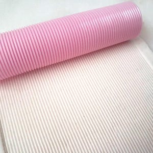 May include: A pink, ribbed rolling pin rests on a white, textured surface. The rolling pin is cylindrical and has a series of parallel ridges along its length. The white surface also has a ribbed texture, creating a pattern of parallel lines.