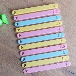 May include: A set of 11 colourful measuring sticks with heart-shaped cutouts at the top. The sticks are yellow, pink, and blue and are labelled with numbers from 1 to 6, with increments of 0.5.