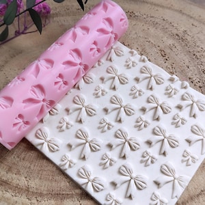 Puede incluir: Rodillo de silicona rosa con un patrón de lazo. El rodillo se utiliza para crear diseños en relieve en fondant o masa. El rodillo se muestra junto a una superficie blanca con un patrón de lazo en relieve.