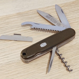May include: An olive green multi-tool pocket knife with a textured handle and a Mauser logo. The knife features multiple deployed stainless steel implements including a saw, two blades, a corkscrew, a small awl, and a separate metal plate.