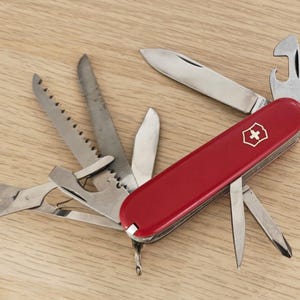 Peut inclure: Un couteau suisse rouge multifonction avec un emblème de croix blanche sur le manche. L'outil est présenté avec plusieurs accessoires en acier inoxydable déployés, dont une lame principale, une scie, des ciseaux et un ouvre-bouteille.