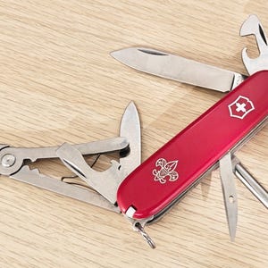 May include: A red multi-tool pocket knife featuring a silver scout emblem and a white cross shield logo. The tool is partially unfolded, revealing a main blade, pliers, a bottle opener, a screwdriver, and a small awl made of stainless steel.