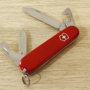 Può includere: Un coltellino svizzero multifunzione rosso con un logo a scudo argentato sul manico. L'utensile include una lama principale, un apribottiglie e una lama più piccola, tutte in acciaio inossidabile, con un anello metallico all'estremità.