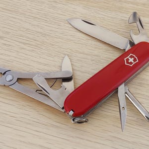 May include: A red multi-tool pocket knife featuring a white Swiss cross emblem on the handle. The tool is fully extended with various stainless steel implements deployed, including pliers, a knife blade, a bottle opener, and a screwdriver.