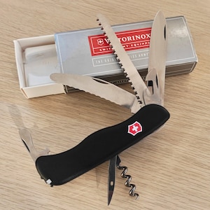 Peut inclure: Un couteau suisse Victorinox noir avec plusieurs outils en acier inoxydable déployés, dont une lame, une scie et un tire-bouchon. L'outil porte le logo au bouclier rouge et blanc et est placé devant sa boîte d'origine argentée.