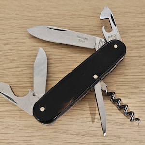 Peut inclure: Un couteau suisse multifonction noir avec plusieurs outils en acier inoxydable déployés, dont deux lames, un décapsuleur, un ouvre-boîte et un tire-bouchon. La lame principale porte l'inscription gravée 'Holliger Bern'.