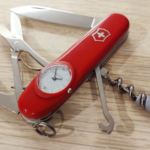 Peut inclure: Un couteau suisse multifonction rouge Victorinox avec un cadran de montre analogique intégré sur le manche. L'outil comprend une lame en acier inoxydable, des ciseaux, un tire-bouchon et un crochet, avec un petit anneau métallique à la base.