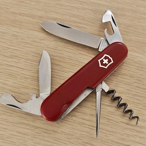Puede incluir: Una navaja suiza Victorinox roja con múltiples herramientas de acero inoxidable desplegadas, incluyendo dos hojas, un abrebotellas, un sacacorchos y un punzón. El mango presenta el icónico emblema de la cruz y el escudo blancos.