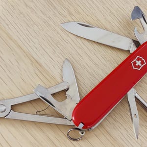 May include: A red multi-tool pocket knife featuring a silver Swiss cross emblem on the handle. The tool is partially unfolded, revealing a main blade, pliers, a bottle opener, a screwdriver, and a small reamer, with a metal key ring at the base.