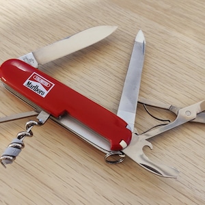 Può includere: Un coltellino svizzero multifunzione rosso con il logo Marlboro Unlimited sul manico. L'utensile è aperto e mostra una lama in acciaio inossidabile, una lima per unghie, forbici piccole, un cavatappi, un apribottiglie e un piccolo gancio.