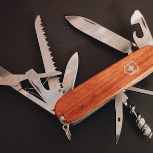 Peut inclure: Un couteau suisse multifonction doté de plaquettes en bois poli avec un emblème argenté en forme de croix et de bouclier. Les outils en acier inoxydable incluent une grande lame, une scie, des ciseaux, un décapsuleur et un tire-bouchon.