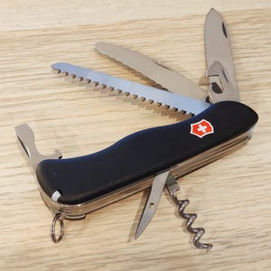 Peut inclure: Un couteau suisse multifonction noir avec un emblème en forme de bouclier rouge et blanc sur le manche. L'outil comprend une lame déployée, une scie, une lame dentelée, un tire-bouchon, un tournevis plat et un petit anneau métallique.