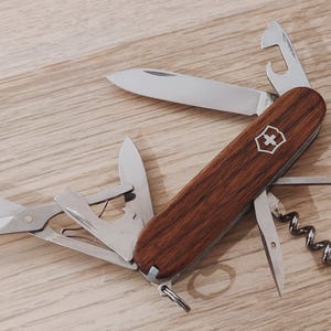 May include: A multi-tool pocket knife with dark brown wood grain scales and a silver cross and shield emblem. The stainless steel tool features an open main blade, scissors, a bottle opener, a corkscrew, a reamer, and a multi-purpose hook.