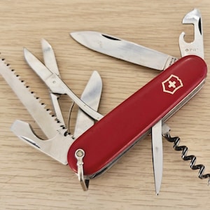Peut inclure: Un couteau suisse multifonction rouge avec un emblème de croix suisse argenté sur le manche. L'outil est entièrement déployé, révélant plusieurs accessoires en acier inoxydable, dont une lame principale, une scie, des ciseaux et un tire-bouchon.