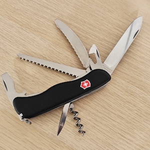 Peut inclure: Un couteau de poche multifonction noir avec un emblème de croix suisse sur le manche. L'outil est partiellement déplié, révélant une lame principale, une lame dentelée, une scie, un décapsuleur, un tournevis et un tire-bouchon en acier.
