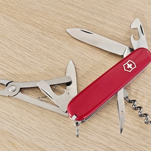 May include: A red multi-tool pocket knife featuring a white cross emblem on the handle. The tool is partially unfolded, revealing a main blade, pliers, a bottle opener, a corkscrew, and a small awl, all constructed from polished stainless steel.