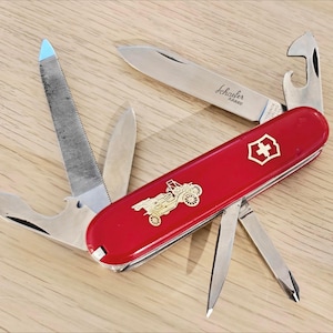 Peut inclure: Un couteau suisse multifonction rouge avec plusieurs outils en acier inoxydable déployés. Le manche présente un emblème doré représentant une calèche et une croix suisse. La lame principale porte l'inscription gravée Schaefer RARE.