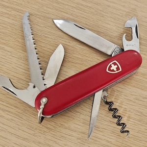 Peut inclure: Un couteau suisse rouge multifonction avec un emblème en forme de bouclier argenté et une croix blanche. L'outil comprend une lame principale, une scie, un décapsuleur, un ouvre-boîte, un tire-bouchon et une petite lame secondaire déployés.