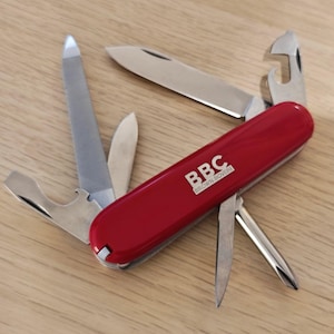 May include: A red multi-tool pocket knife with several stainless steel implements extended, including a blade, file, screwdriver, and bottle opener. The handle features the white printed text BBC BROWN BOVERI on the side of the casing.