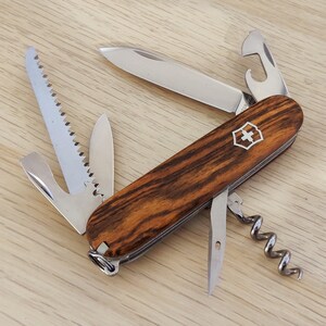 Peut inclure: Un couteau de poche multifonction avec des plaquettes en bois brun foncé veiné et un logo bouclier argenté. L'outil comprend une lame principale, une scie, un décapsuleur, un tire-bouchon et un poinçon en acier inoxydable poli.
