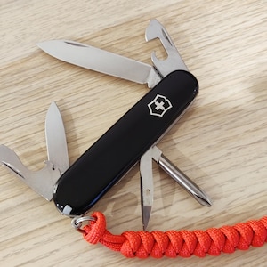 Peut inclure: Un couteau de poche multifonction noir avec un emblème argenté, comprenant une grande lame, une petite lame, un décapsuleur, un tournevis et un poinçon. Une dragonne en paracorde tressée orange vif est fixée à l'anneau métallique à la base.