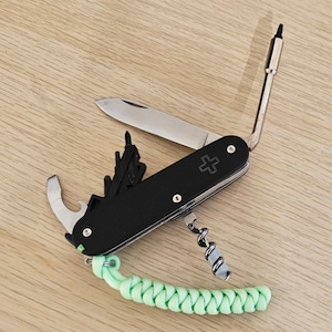 May include: A black multi-tool pocket knife featuring a silver blade, a bottle opener, a corkscrew, and an extended screwdriver bit. The handle has a textured black grip with a cross emblem and a light green braided paracord lanyard attached.