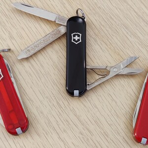 Peut inclure: Trois outils multifonctions avec un emblème de croix suisse. Un outil noir est ouvert, révélant une lame, une lime à ongles et des ciseaux. Deux outils rouges sont fermés, l'un translucide et l'autre opaque brillant, avec un anneau métallique.