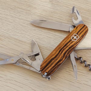 Peut inclure: Un couteau de poche multifonction doté d'un manche en bois veiné avec un emblème argenté en forme de croix et de bouclier. Les outils en acier inoxydable incluent une lame, des ciseaux, un tire-bouchon et un décapsuleur déployés.