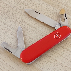 Peut inclure: Un couteau de poche multifonction rouge avec un emblème en forme de bouclier blanc orné d'une croix. L'outil présente deux lames en acier inoxydable déployées, l'une avec un tournevis plat et un décapsuleur, l'autre avec une lame et un ouvre-boîte.