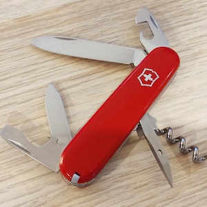 Può includere: Un coltellino svizzero rosso multifunzione con un emblema a croce bianca sul manico. L'utensile presenta diversi componenti in acciaio inossidabile aperti, tra cui una lama principale, una lama piccola, un cavatappi e un apribottiglie.