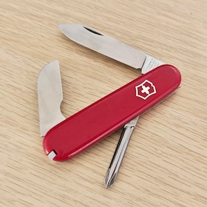 Peut inclure: Un couteau de poche multifonction rouge avec un emblème argenté en forme de croix et de bouclier sur le manche. L'outil présente trois ustensiles en acier inoxydable déployés : une grande lame, une petite lame et un tournevis cruciforme.