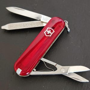 Peut inclure: Un couteau suisse rouge avec un manche translucide et un emblème de croix argenté. L'outil multifonction présente une lame en acier inoxydable déployée, une lime à ongles avec un bout plat et des ciseaux à ressort déployés sur le côté.