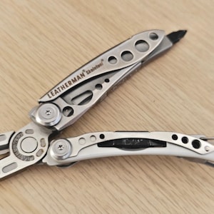 Könnte beinhalten: Ein silbernes Leatherman Skeletool Multifunktionswerkzeug mit Spitzzange, Drahtschneider und Schraubendreher-Bit. Die Metallgriffe haben ein skelettiertes Design mit kreisförmigen Aussparungen und einem integrierten Karabinerhaken.